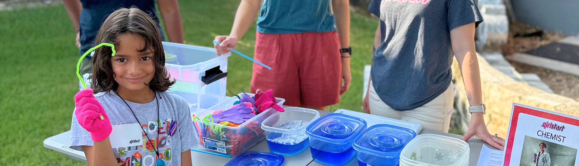 A Girlstart participant stands in front of an activity station in Girlstart's backyard. She is smiling and holding a green pipe cleaner.